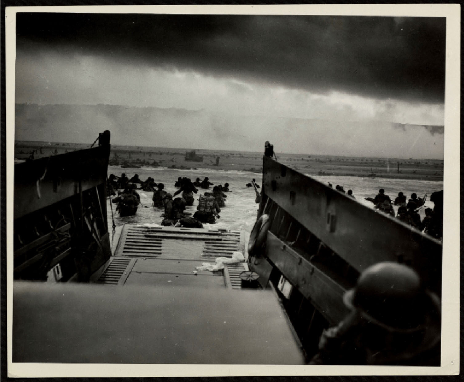View from inside a landing craft as soldiers disembark onto a sandy beach under an overcast sky during a historic military operation