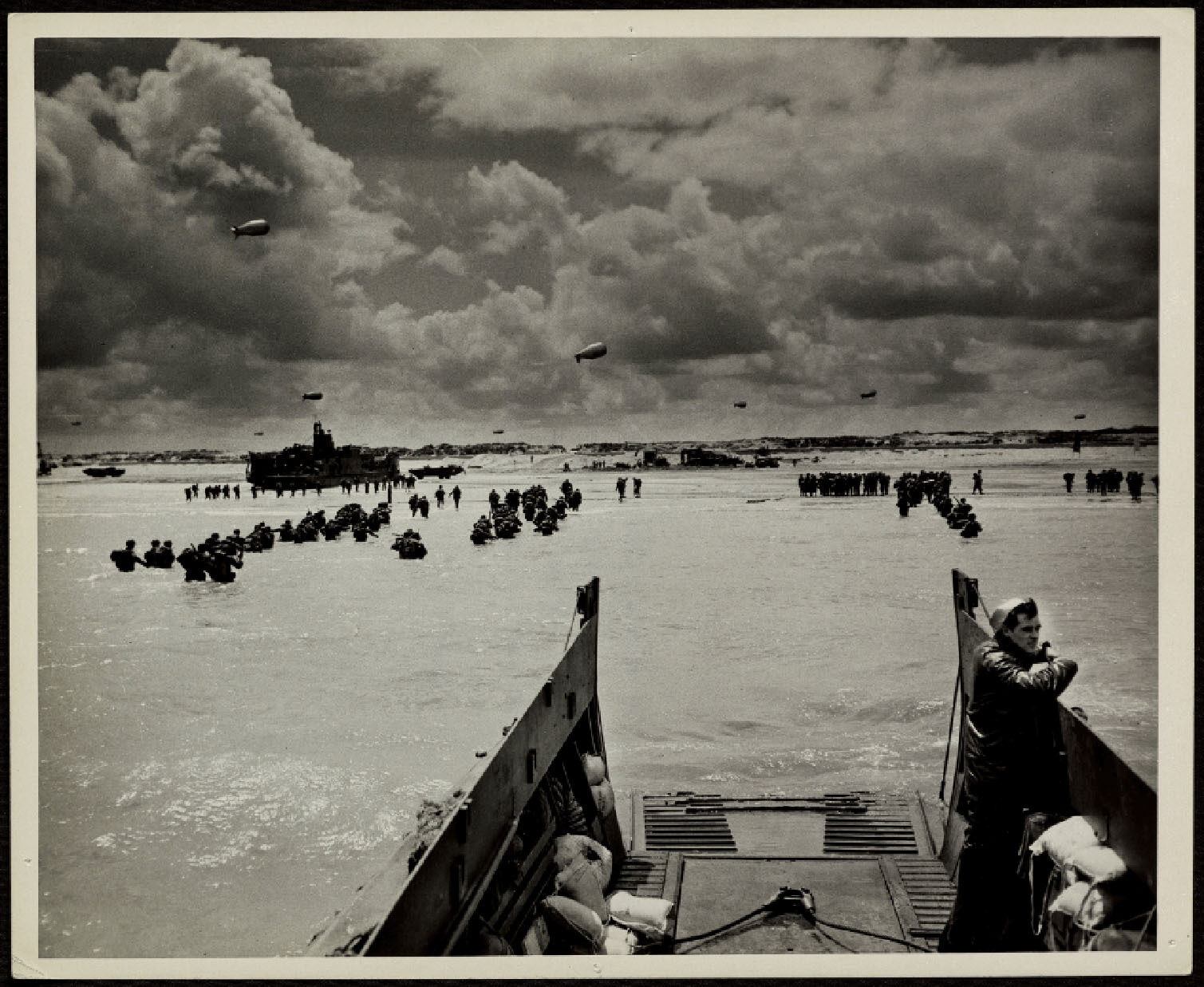 View from a landing craft as soldiers wade through shallow water towards a beach during D-Day, with parachutes and aircraft in the cloudy sky
