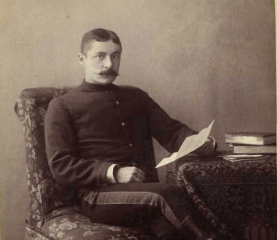 A man in a vintage military uniform sits on a chair, holding a letter with books on a table.