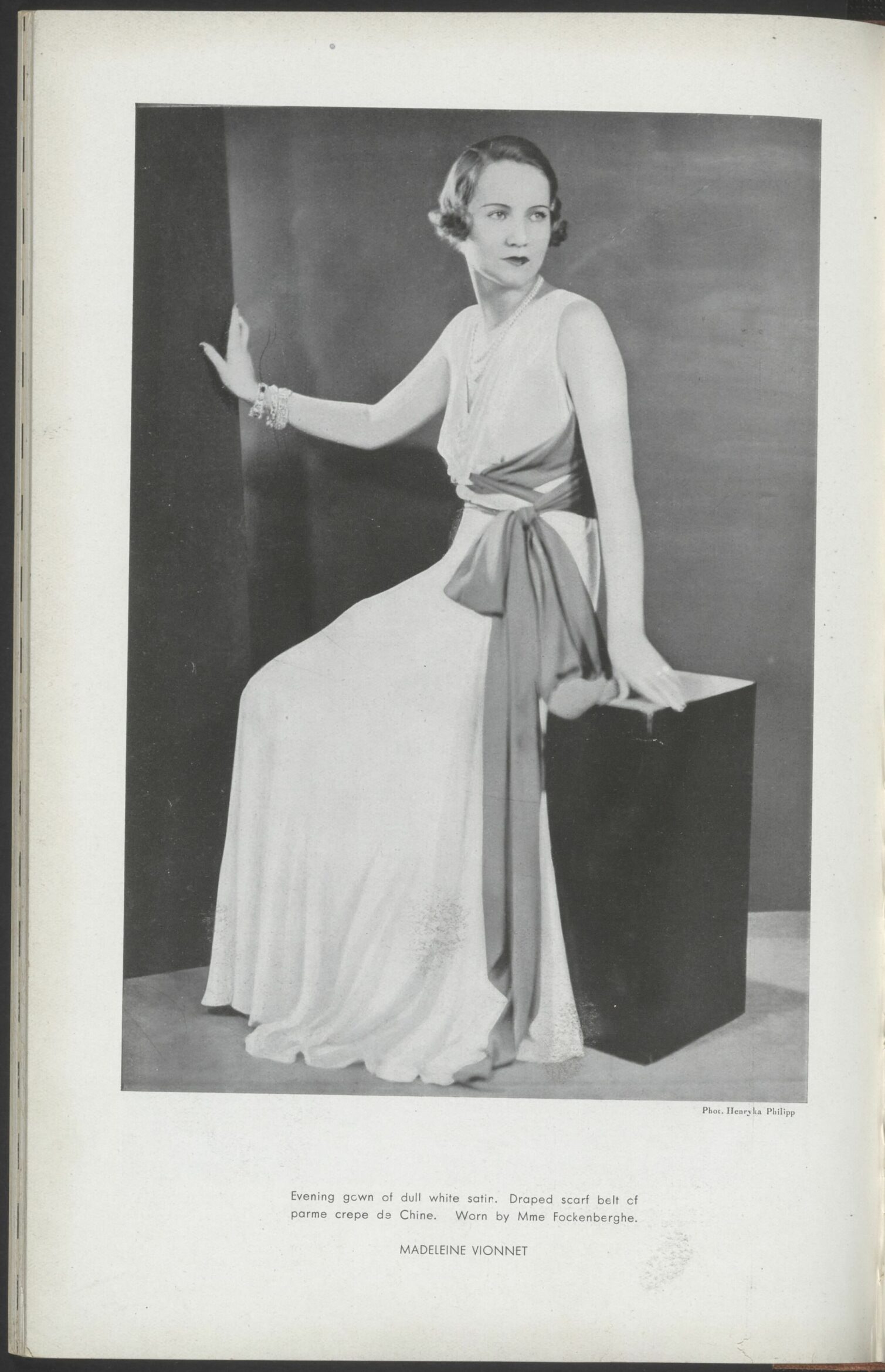 A woman in a flowing white dress with a sash, sitting on a box and posing gracefully.