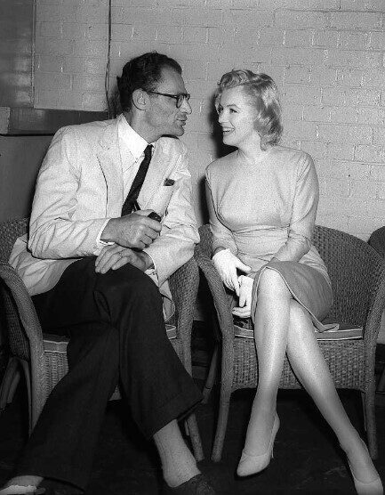 A man in a light-coloured suit and glasses talks to a woman in a light dress with a smile, sitting in wicker chairs.
