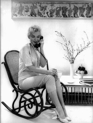 A woman sitting on a rocking chair, talking on a phone, holding a cup in hand.