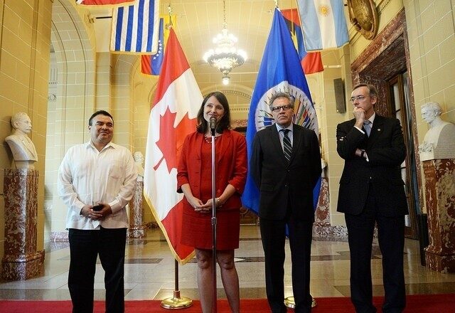 Four people standing in a hall with flags, one person speaking into a microphone.