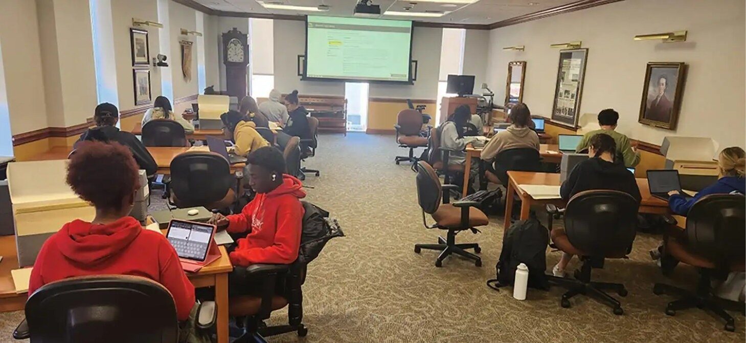 Students working at desks with laptops and books in a study room.