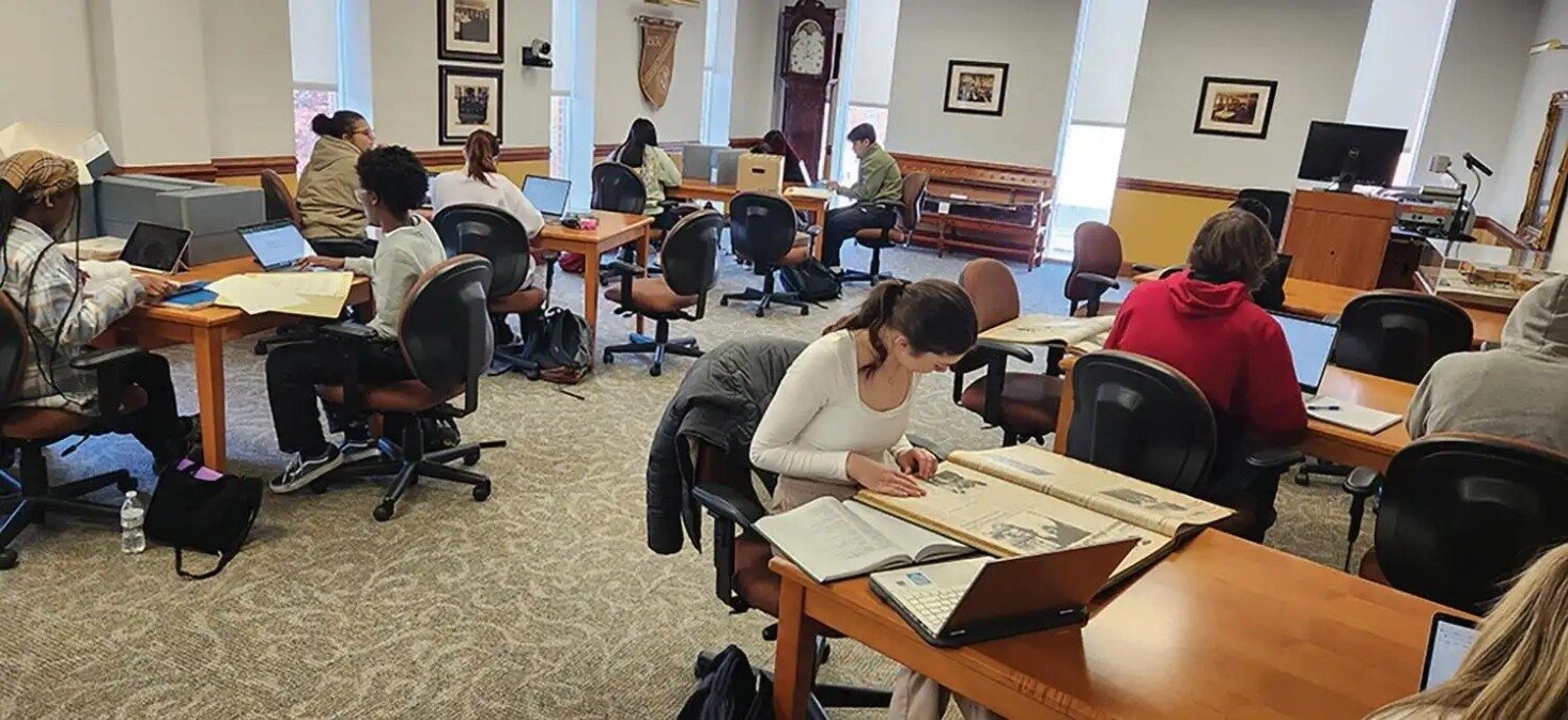 Students working at desks with laptops and books in a study room.
