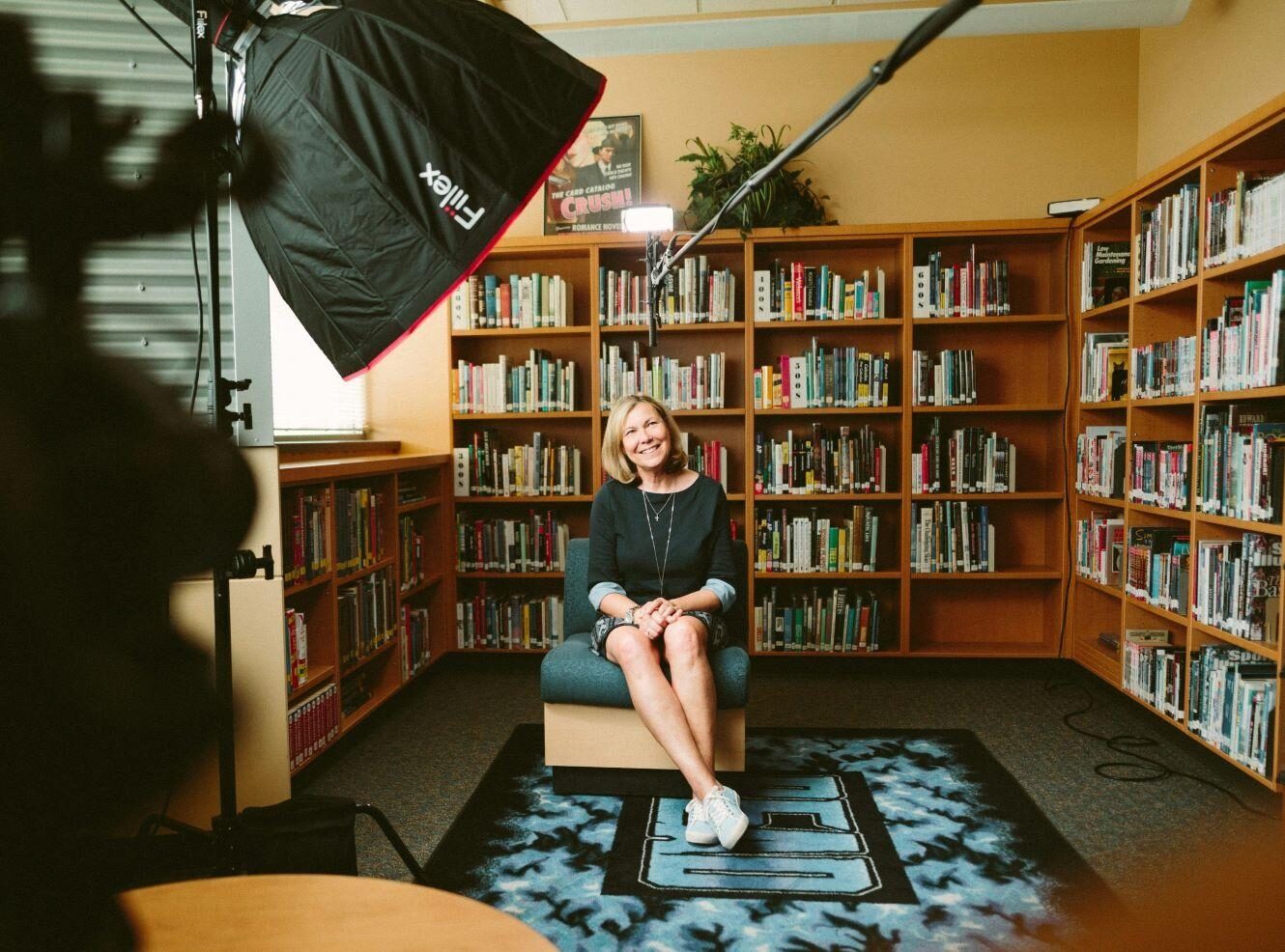 A woman sitting on a chair in a library, smiling. Bookshelves filled with books are in the background.