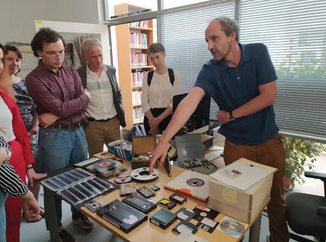 A group of people gathered around a table, examining old media items like tapes and discs.