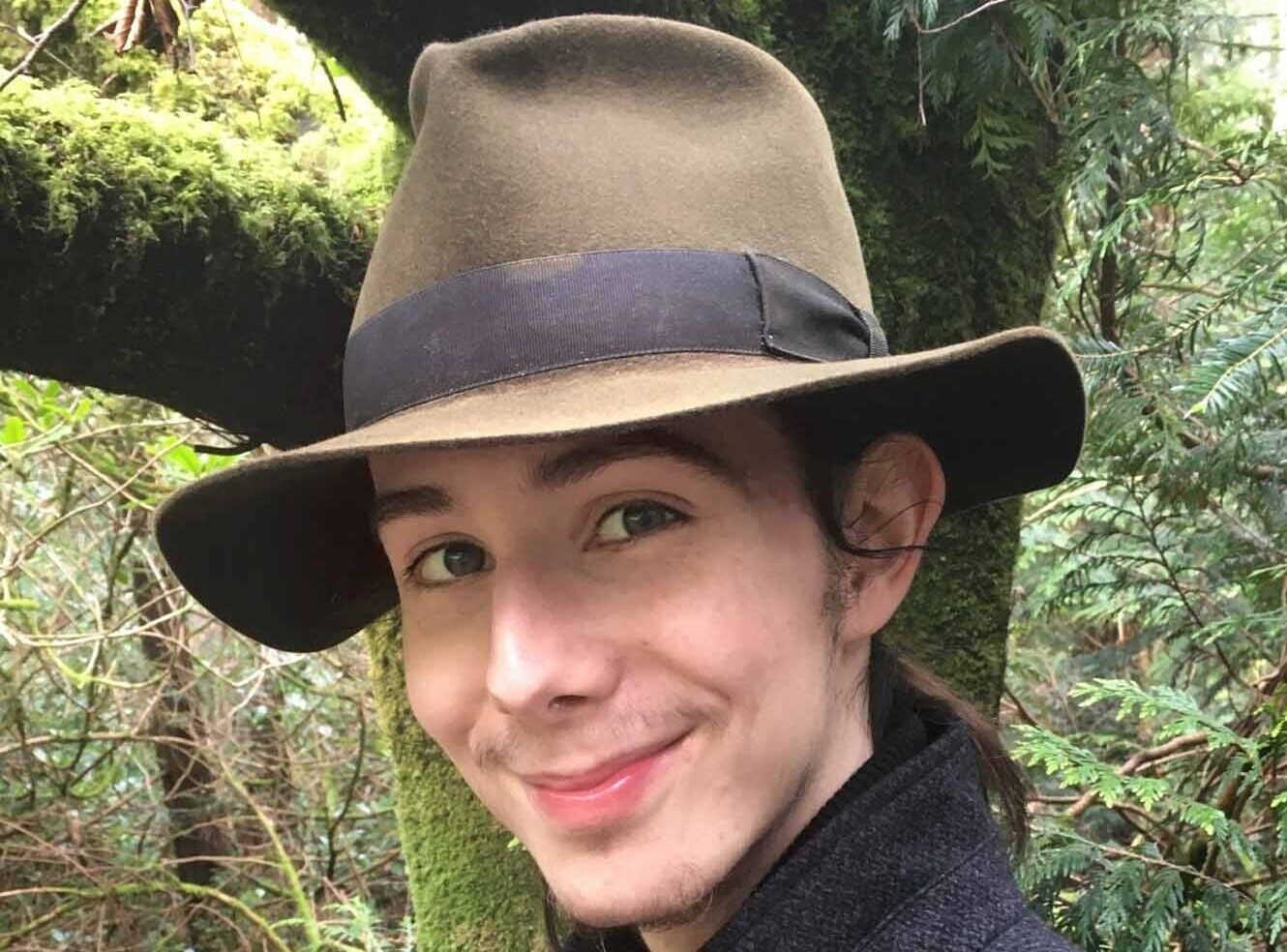 A young person wearing a brown fedora hat smiles in a forest setting.