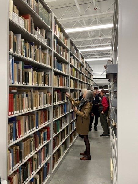 A woman browsing books on a library shelf while others stand nearby.
