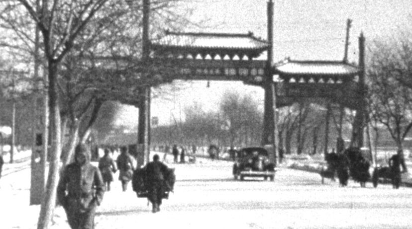 A snowy street with pedestrians in winter clothing walking alongside barren trees, a vehicle driving, and a traditional gate spanning the road.
