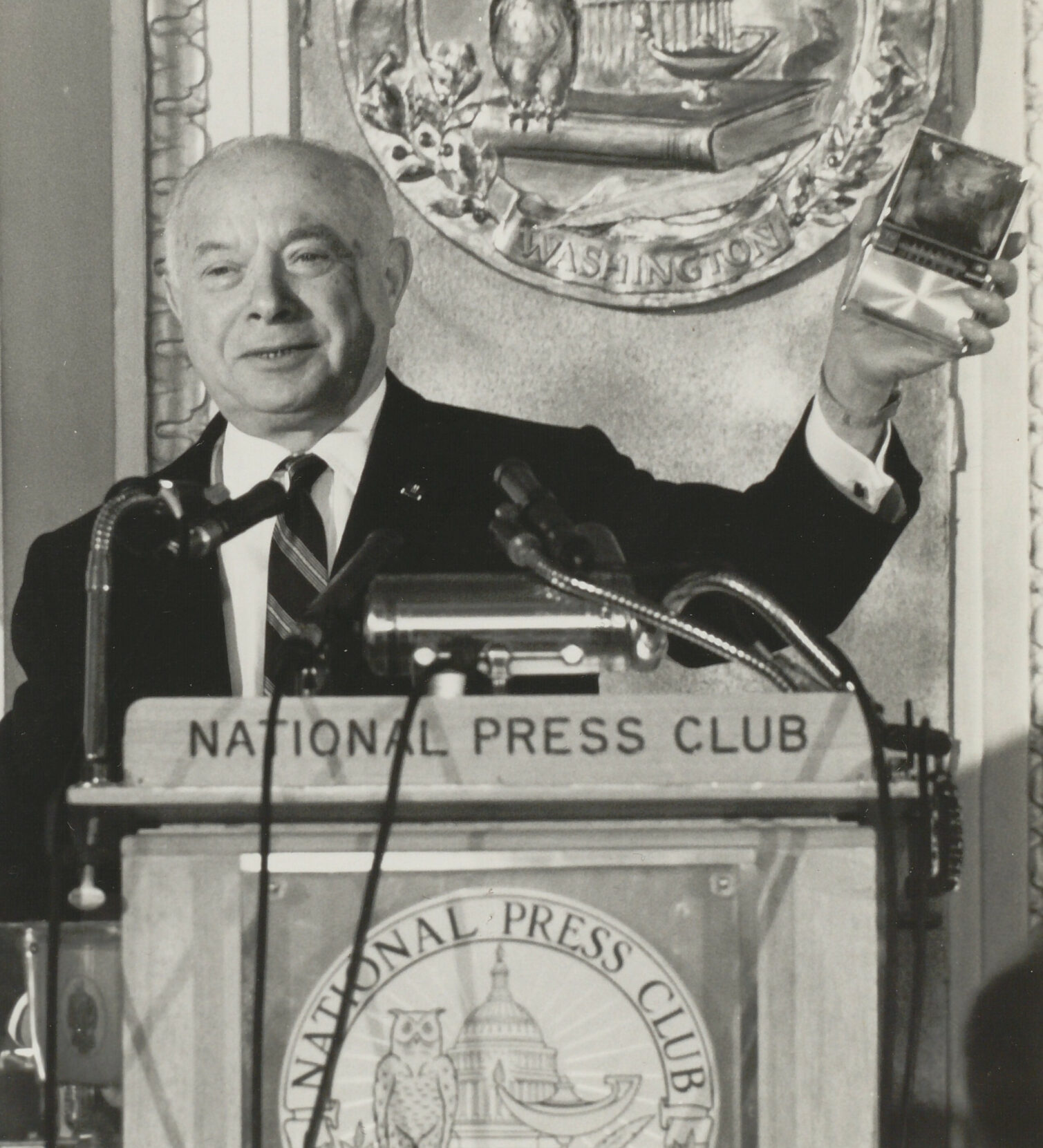 A man speaking at a podium with a sign for the National Press Club, holding a clear award