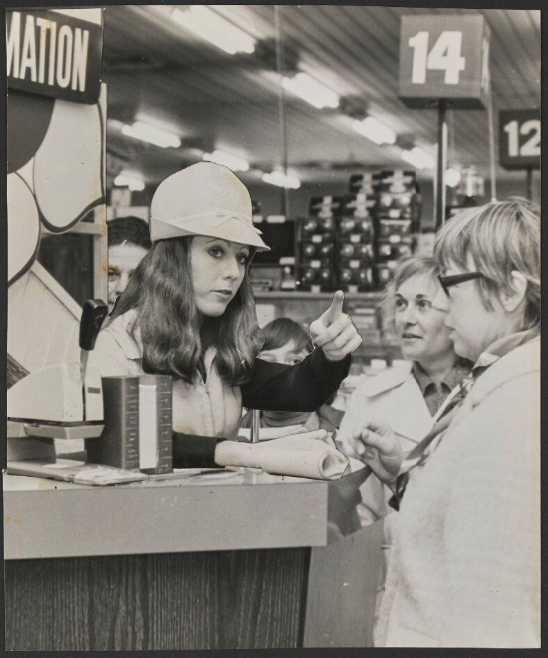 A woman with long hair in a white cap gestures while speaking to three customers at a service counter.