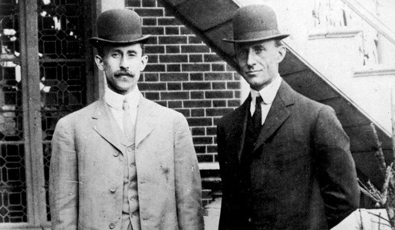 Two men in three-piece suits and bowler hats in front of a brick building.