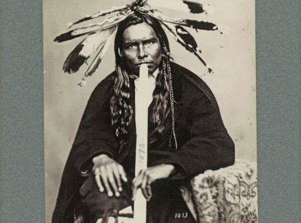 A man wearing a feather headdress plays a long flute, seated with hands on knees.