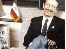 A man in a suit sits in a chair, smiling, with a flag behind him and a photo nearby.
