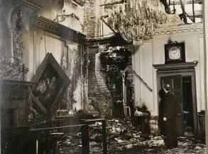A damaged room with a chandelier, burnt walls, and debris scattered on the floor.