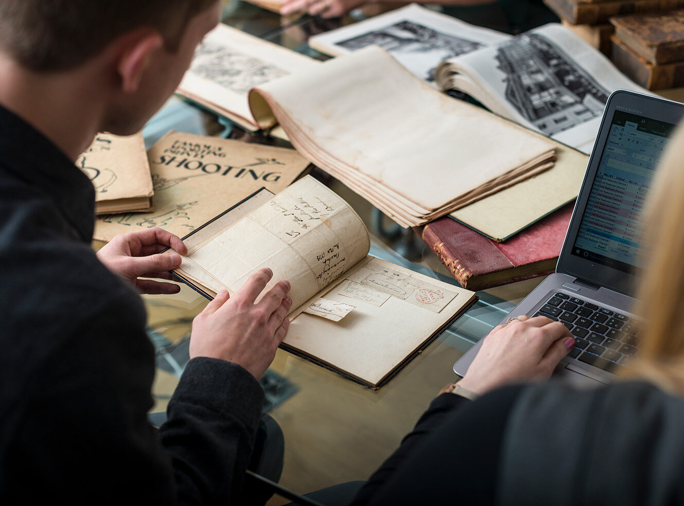 Two people reviewing old documents and books on a table with a laptop.