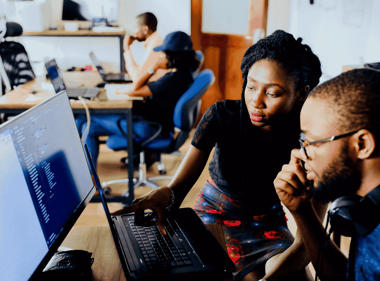 Two people on a laptop and monitor, with one pointing at the screen. Others are working in the background.