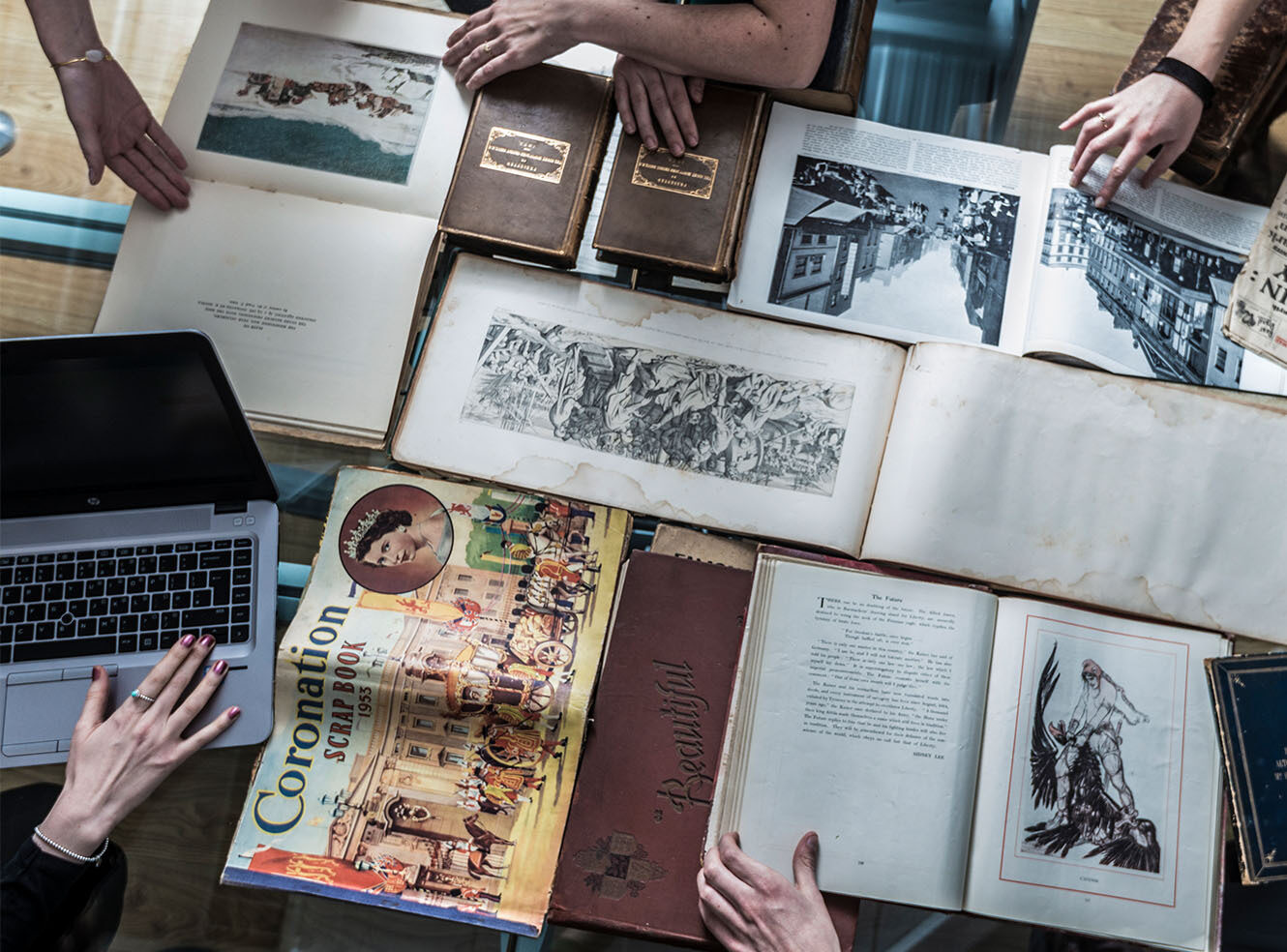 Hands examining antique books, prints, and a newspaper spread across a table, with a laptop nearby.