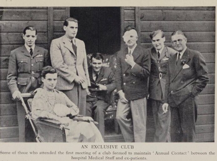 A group of seven men pose together outside a building, with one man seated in a chair