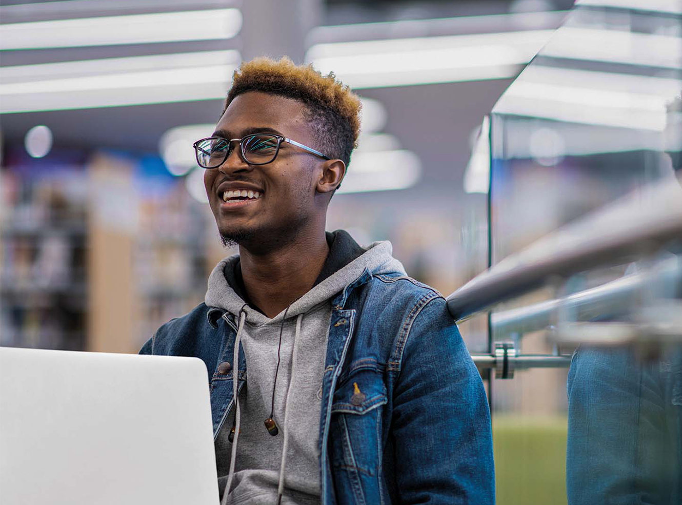 A man in a denim jacket smiling while working on a laptop in a library