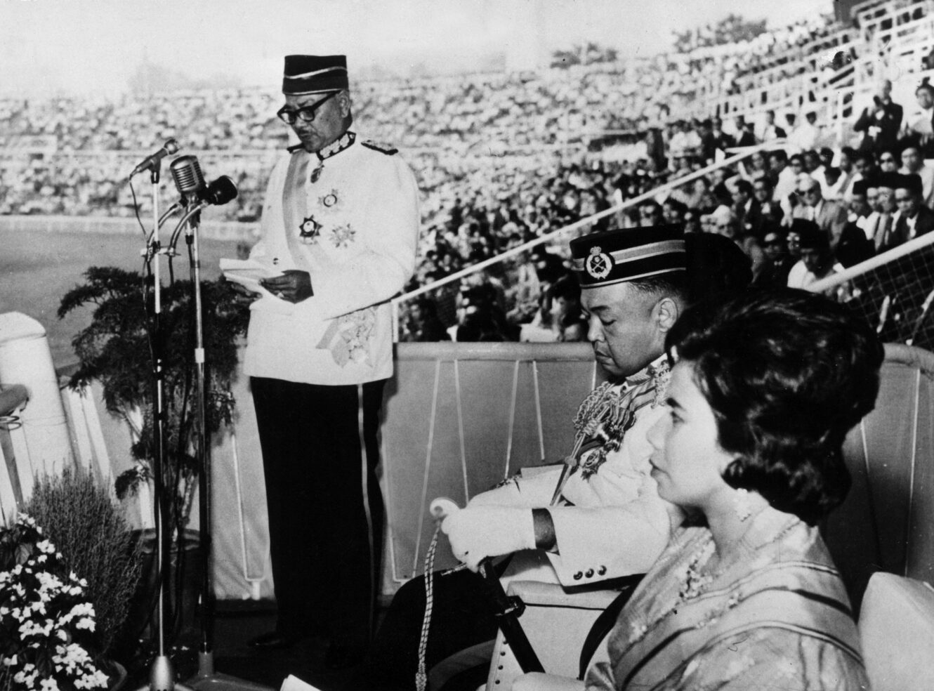 A black and white photo of a man in military uniform giving a speech on stage, while a woman in traditional attire and another man in uniform look on, with an audience in the background