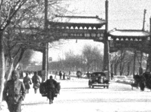 A snowy street with pedestrians in winter clothing walking alongside barren trees, a vehicle driving, and a traditional gate spanning the road.