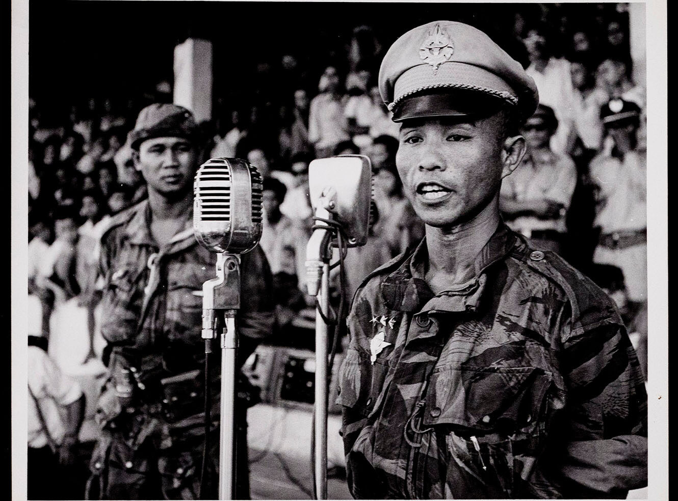 Two soldiers in military uniforms standing at microphones, with a crowd in the background