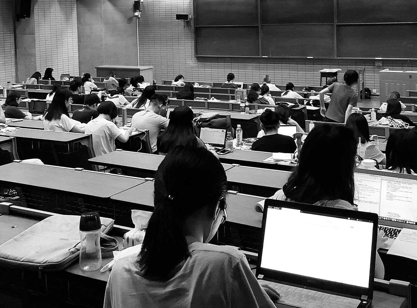 A classroom filled with students studying at desks, some using laptops, in black and white.