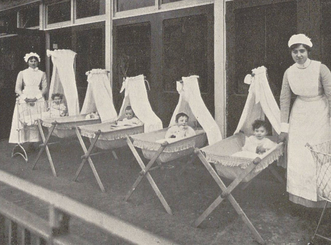 Two women in white nurse uniforms stand by cradles with infants cradles, outdoors.