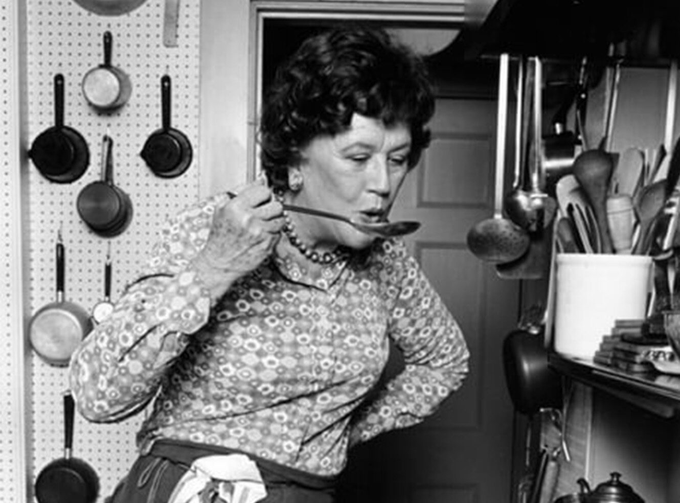 A woman in a kitchen tasting food from a ladle. Cooking utensils and pans surround her.