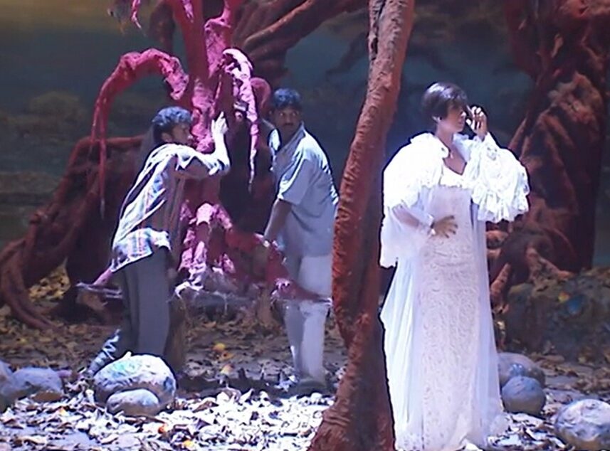 Two actors dressed in gray and white perform on stage beside a red tree, with a third actor in a long white dress