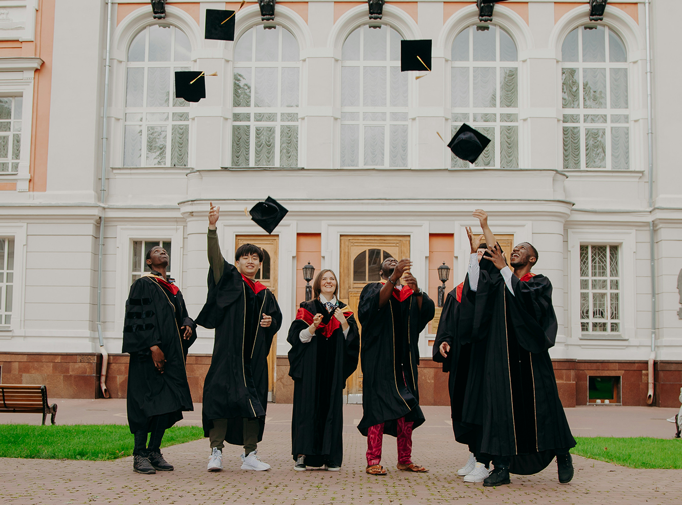 Graduates in caps and gowns toss their hats in front of a university building