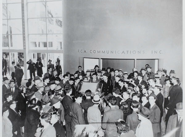 A large crowd of people gathered around a display at RCA Communications, dressed in 1930s fashion.