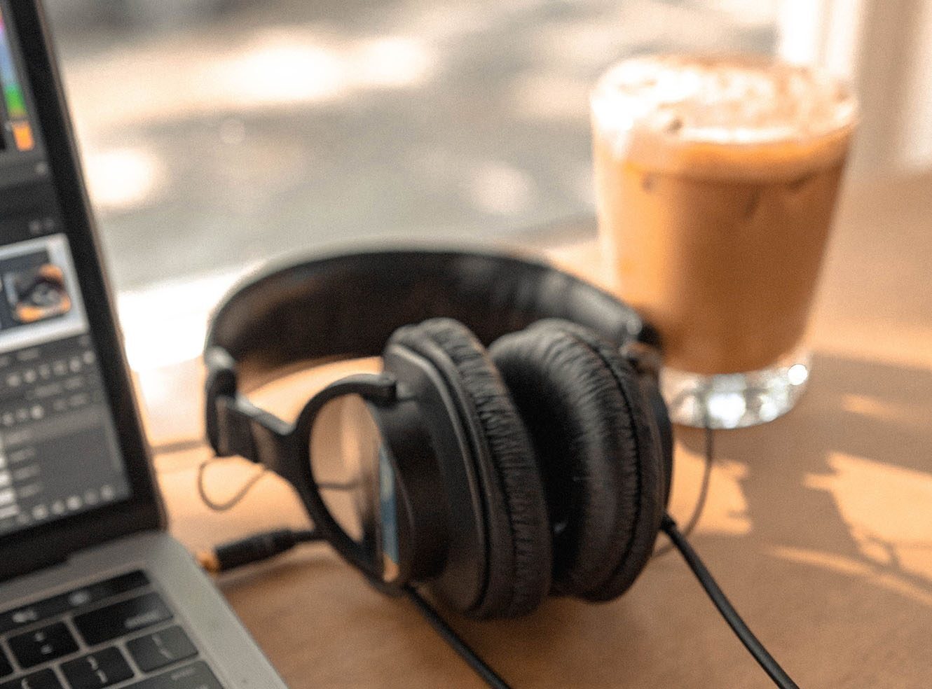 Black overhead headphones plugged into a laptop on a brown table next to a glass of iced coffee.