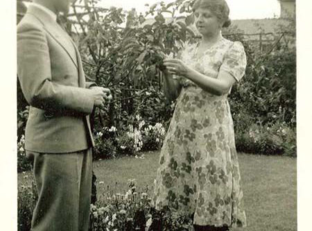 A man in a suit and a woman in a floral dress stand in a garden, discussing a small plant.