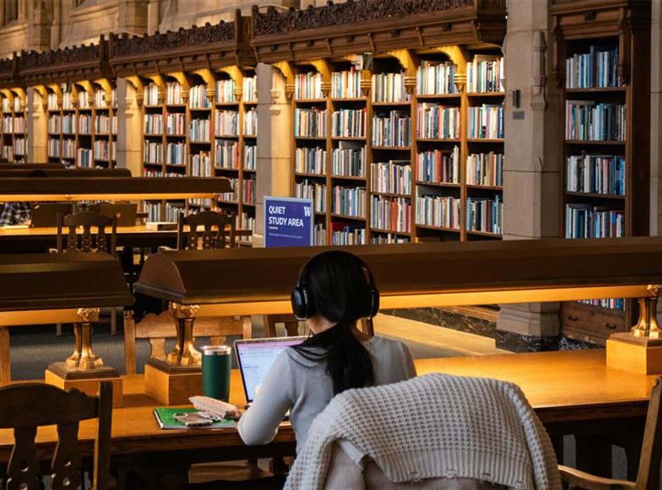 A student with headphones studying at a wooden table in a library filled with books.