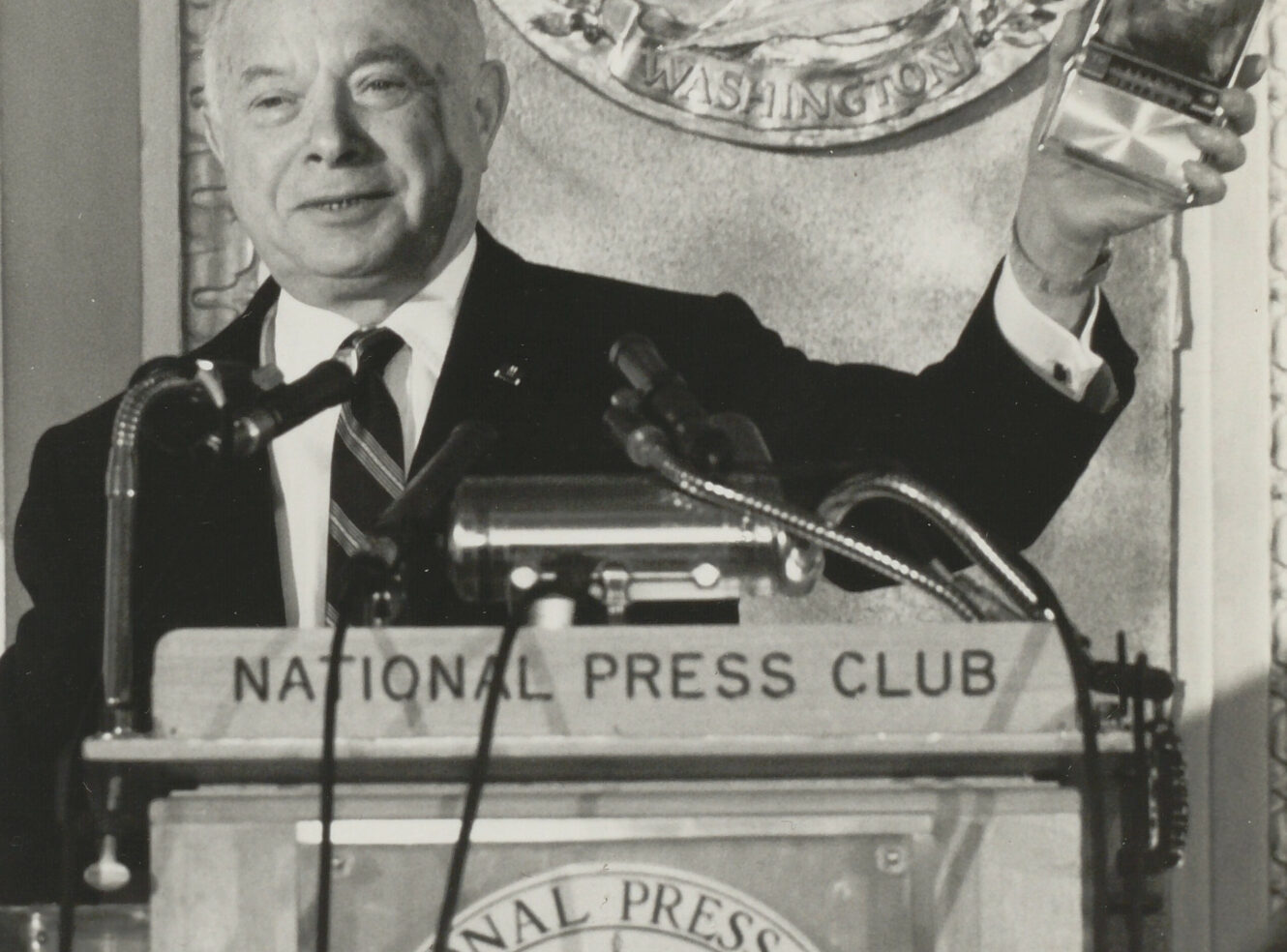 A man speaking at a podium with a sign for the National Press Club, holding a clear award