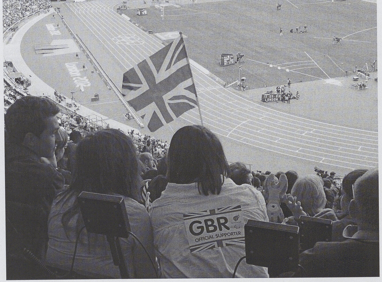 Two people in the stands at a track event, one holding a British flag and wearing a GBR jacket.