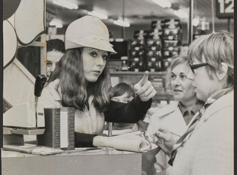 A woman with long hair in a white cap gestures while speaking to three customers at a service counter.