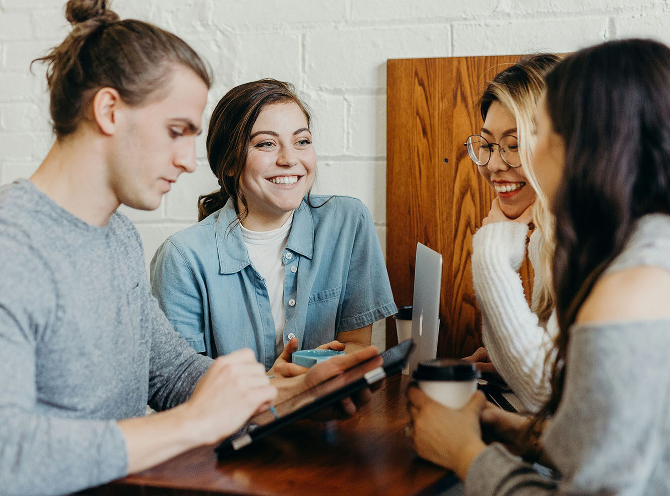 Four young adults at a table, smiling and collaborating with a tablet, laptop, and coffee cup.