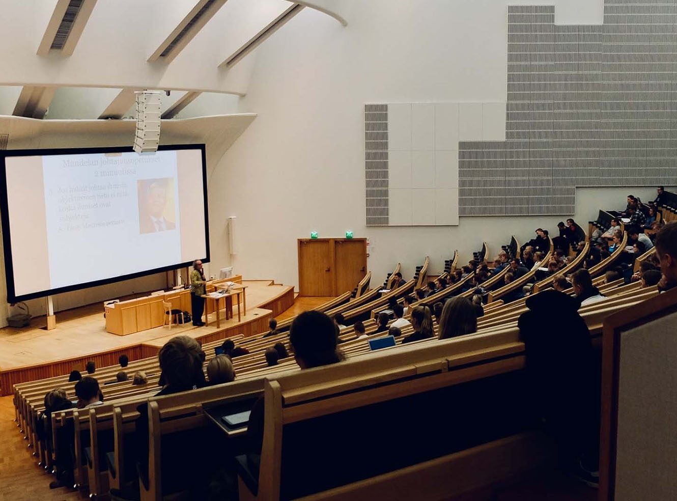 A lecture hall with students sitting in rows, a speaker presenting near a screen with text and an image.