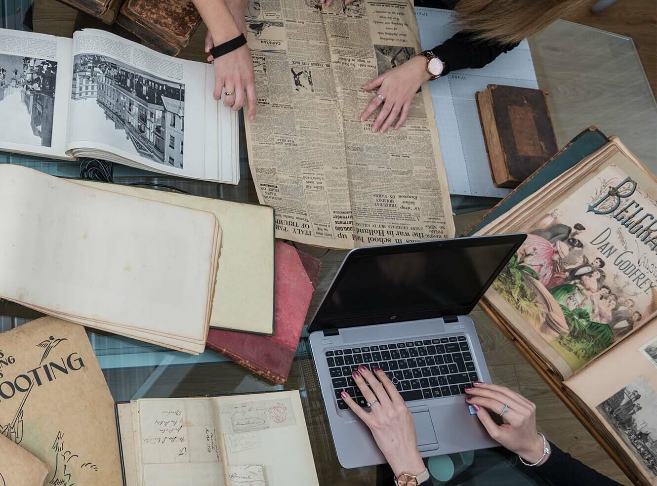 Two people studying historical documents on a glass table, including open books, a newspaper, and a laptop, blending modern tech with vintage research