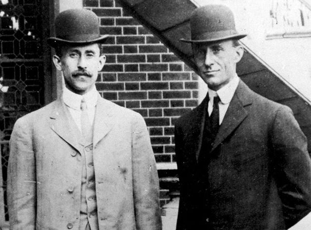 Two men in three-piece suits and bowler hats in front of a brick building.