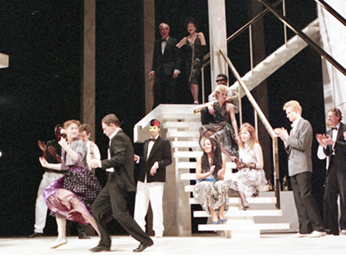 A lively stage scene with dancers and audience clapping, set on a grand staircase