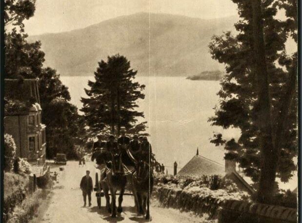 A scenic view of a lake surrounded by trees with two horses and people walking on a road.
