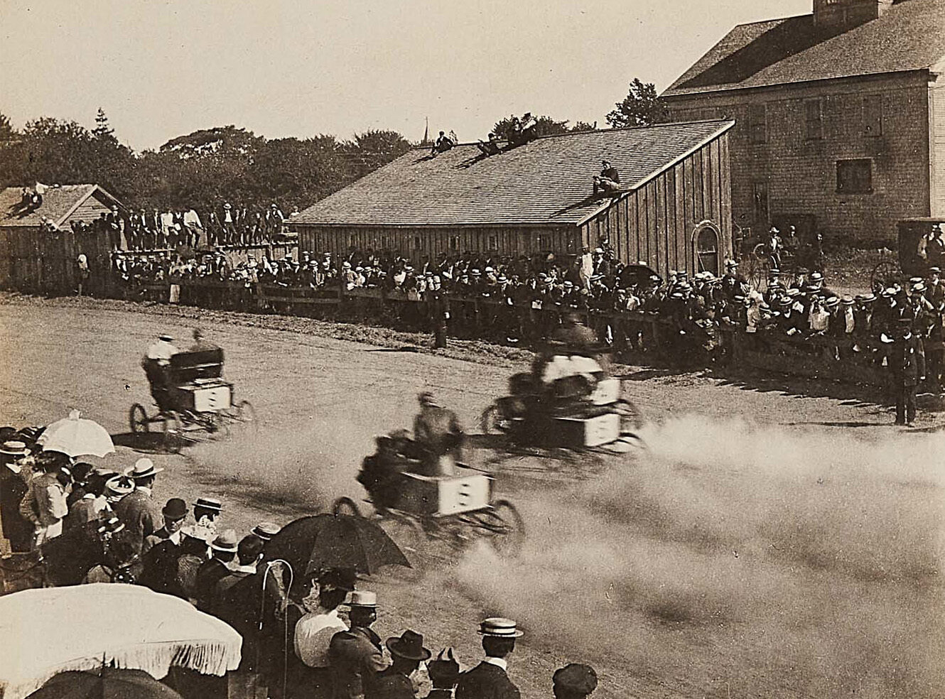 An early motor car race with 4 cars racing and spectators on both sides of the track.