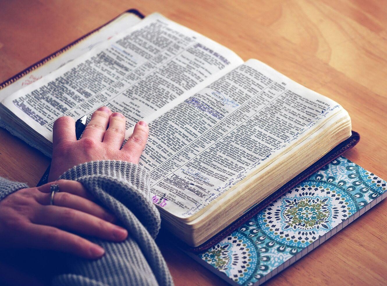 A hand resting on an open Bible with handwritten notes, next to a colourful notebook.