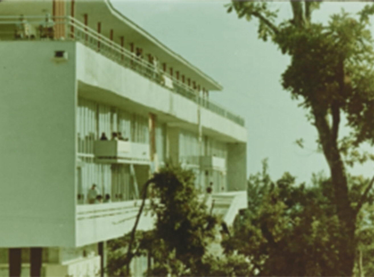 A modern building with multiple balconies surrounded by green trees, under a sunny sky.