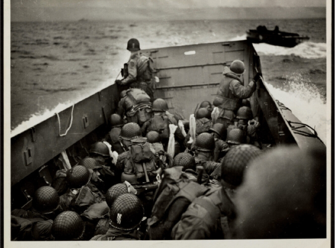 Soldiers in helmets and military gear sit and stand inside a landing craft at sea with another craft visible in the background under an overcast sky